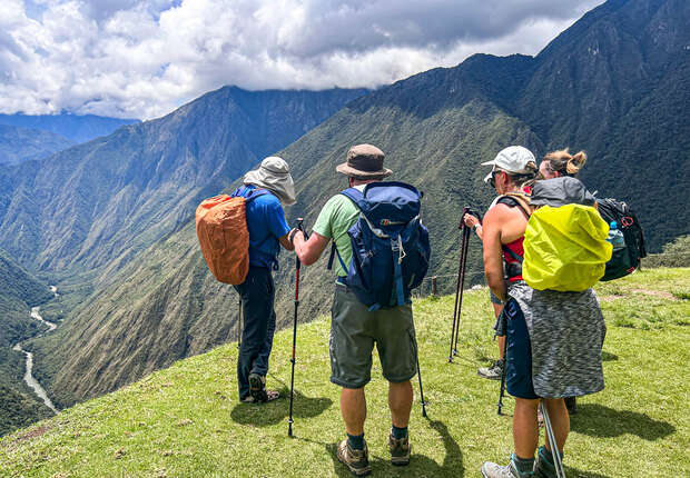 Hikers rest with trekking poles on a grassy mountain ridge, overlooking a lush valley with winding river; dramatic peaks rise under a partly cloudy sky.