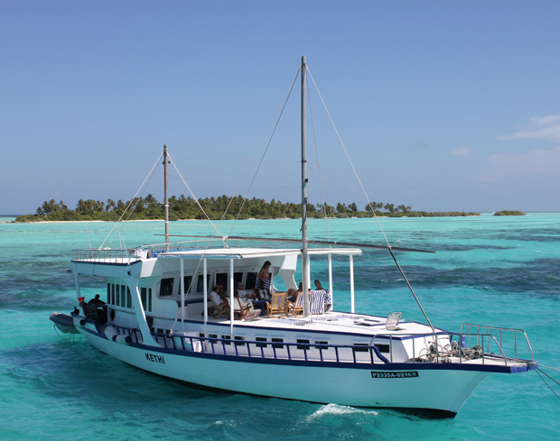 A white boat named "Kethi" floats on turquoise water, carrying passengers seated under a canopy. An idyllic island with lush greenery is visible in the background.