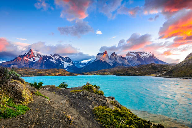 Snow-capped mountains rise majestically, framing a vivid turquoise lake, with rugged terrain and vibrant greenery in the foreground, under a dramatic, colorful sky at sunset.