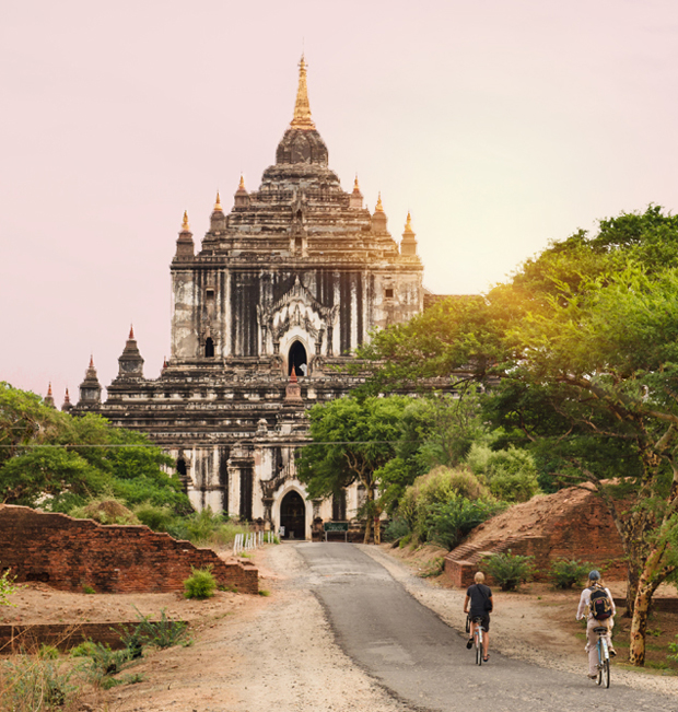 A historical temple stands tall, basking in the warm sunset. Two cyclists ride on a winding path toward it, surrounded by lush greenery and ancient brick ruins.