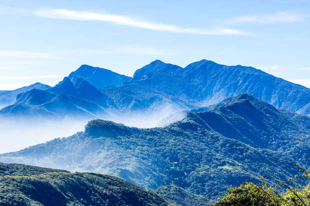 Mountains rise with lush greenery, layered under a clear blue sky, partially obscured by mist drifting across the midsection, creating a serene and expansive natural landscape.