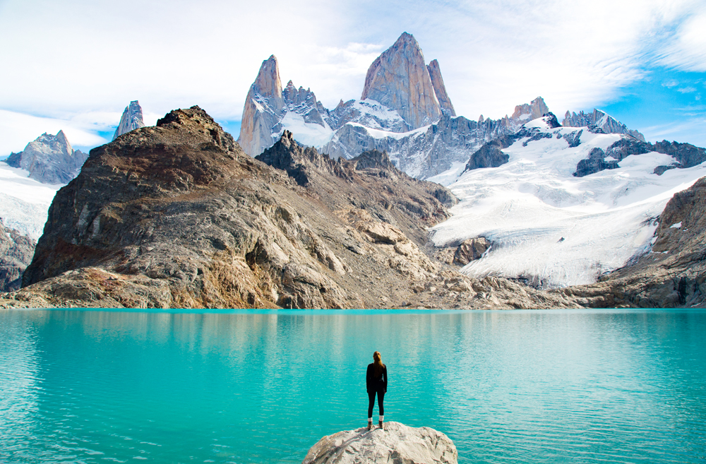 A person stands on a rock by a turquoise lake, gazing at rocky mountains with snow-capped peaks under a partly cloudy sky.