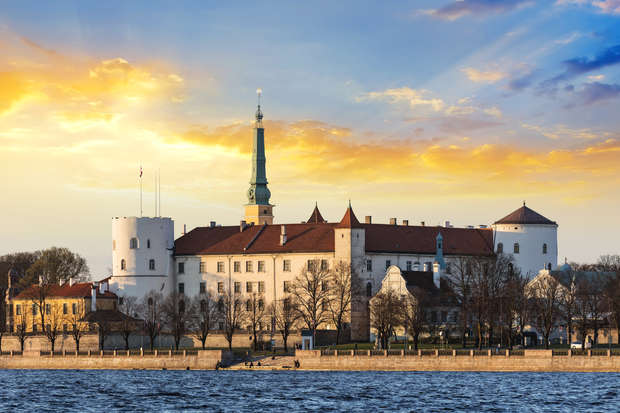 A large, historic building with a prominent spire and red-tiled roof sits beside a river, framed by trees. The sky behind showcases a vibrant sunset with orange and blue hues.