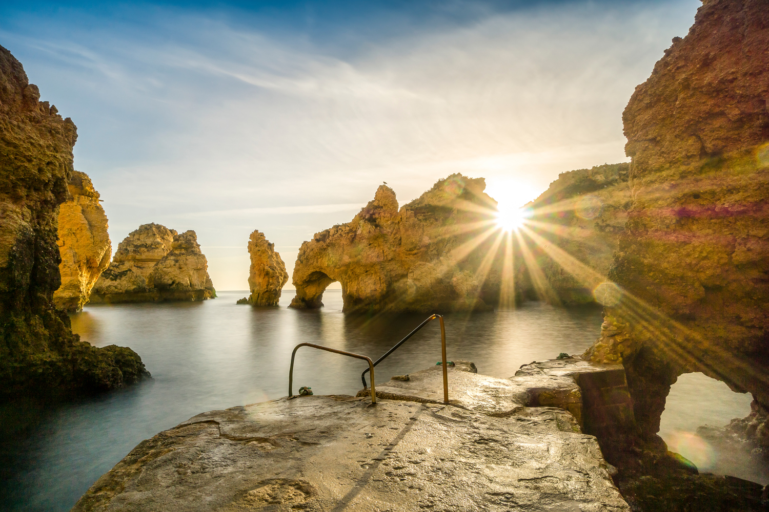 Rock formations rise from the ocean, with sunlight streaming through gaps, near a rough stone platform featuring a metal railing, under a partly cloudy sky.