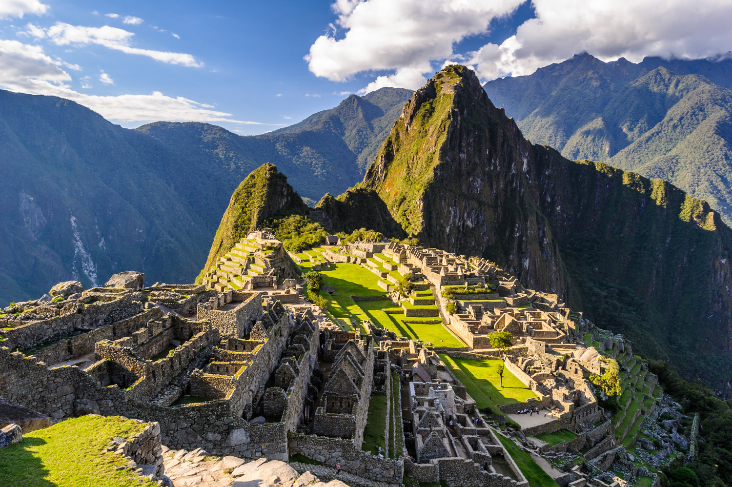 Ancient stone ruins of Machu Picchu rest atop a lush mountain ridge, surrounded by green peaks and under a blue sky with scattered clouds.
