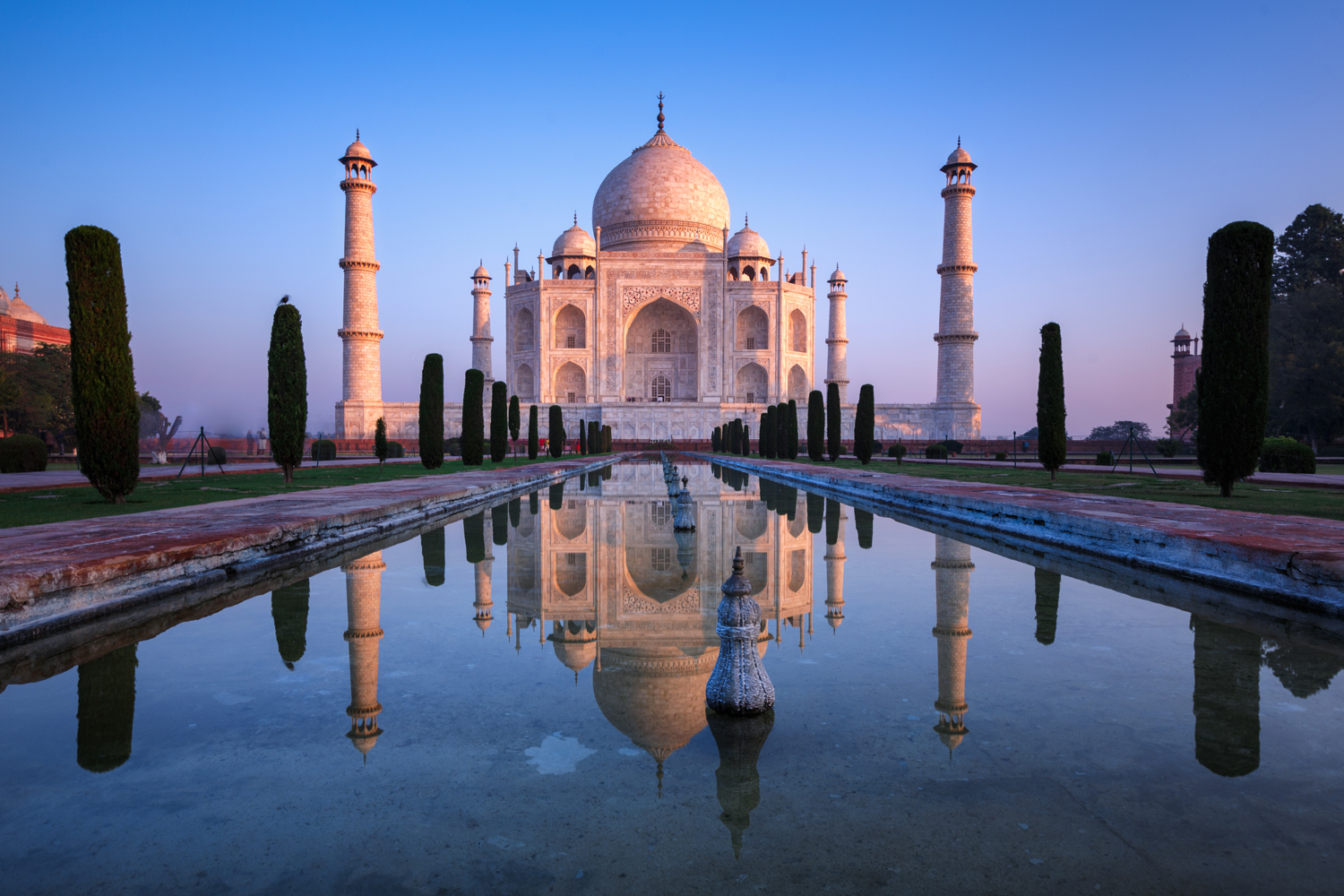 The Taj Mahal stands majestically reflected in a still rectangular pool, surrounded by lush gardens and flanked by tall minarets under a clear blue sky.