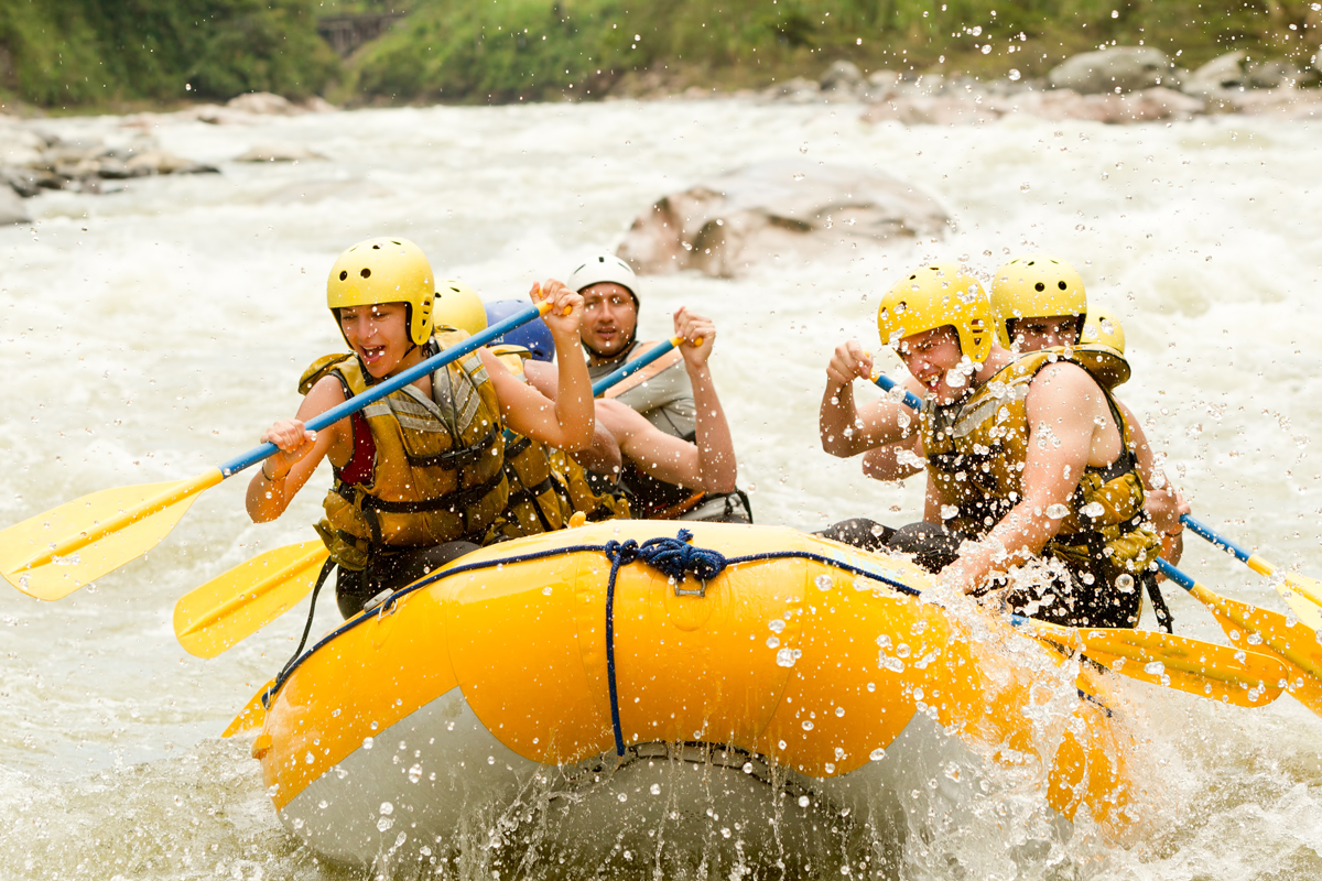 A yellow raft navigates through turbulent white-water rapids, with four paddlers in helmets and life jackets energetically rowing, surrounded by splashing water and rocky riverbanks.
