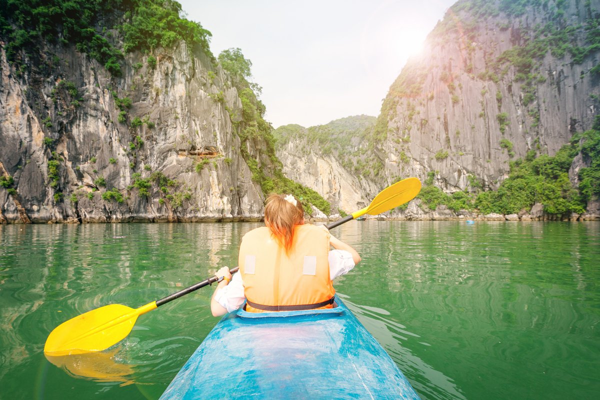 A person in a life vest paddles a blue kayak with a yellow paddle through a narrow waterway, surrounded by towering rock cliffs and lush greenery under bright sunlight.