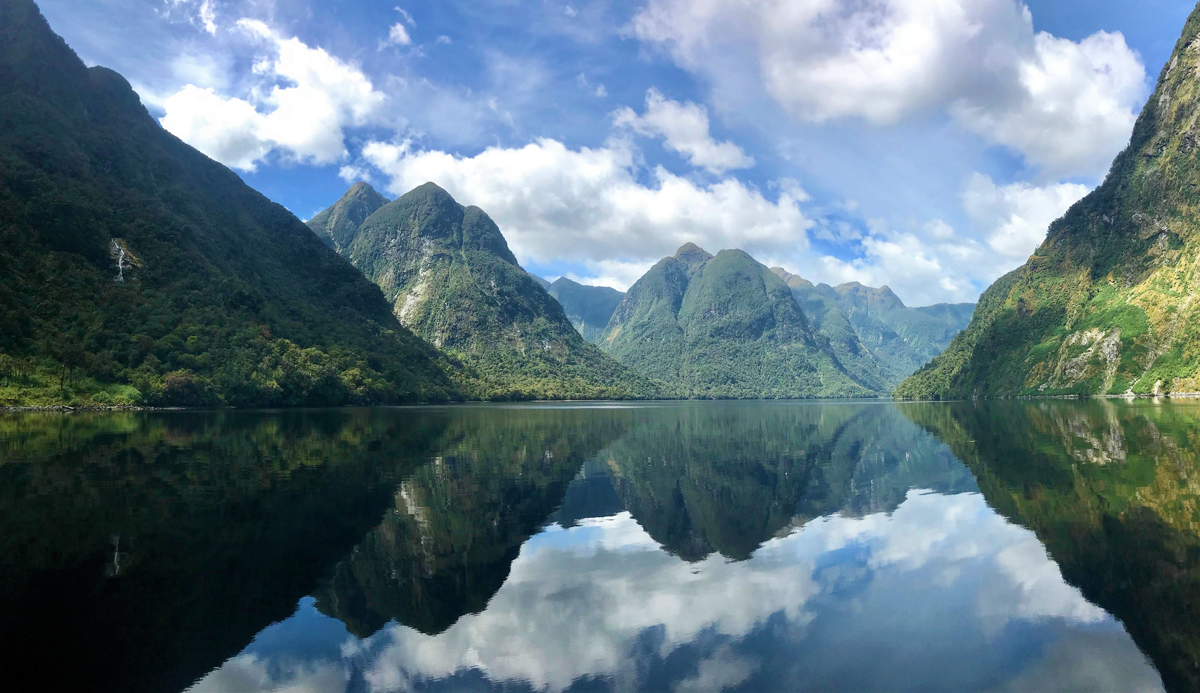 Mountains reflect in a calm lake, surrounded by lush greenery and under a partly cloudy blue sky, creating a serene, symmetrical landscape view.
