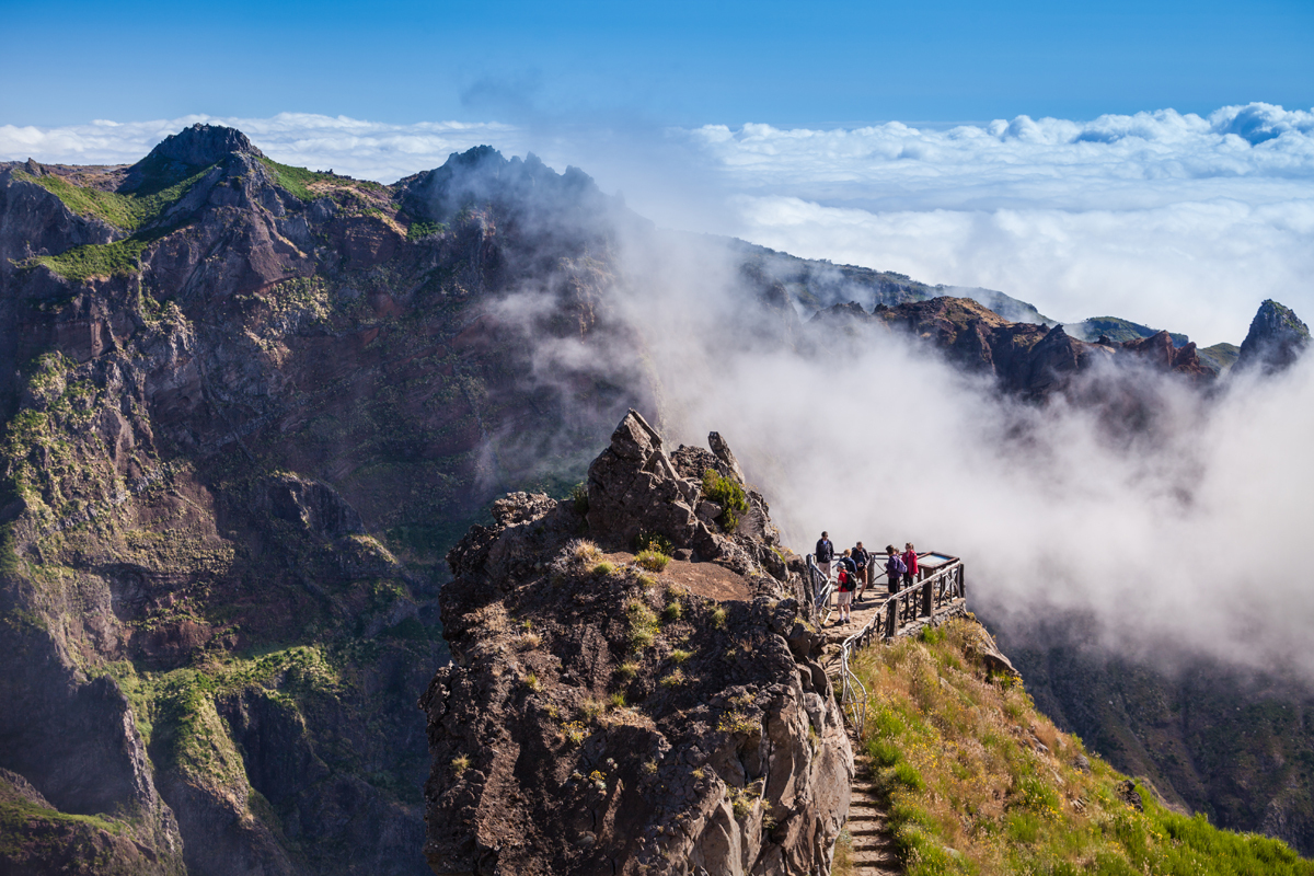 Rocky mountain peak with people walking on a path, surrounded by clouds and fog. Green vegetation contrasts with the rugged rocks, under a clear blue sky.