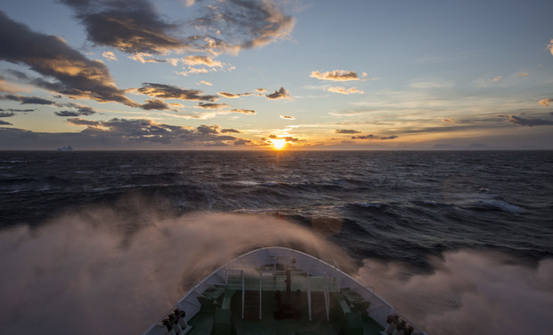 A ship's bow slices through choppy ocean waters, creating misty spray under a vibrant sunset sky, with scattered clouds and the sun near the horizon.