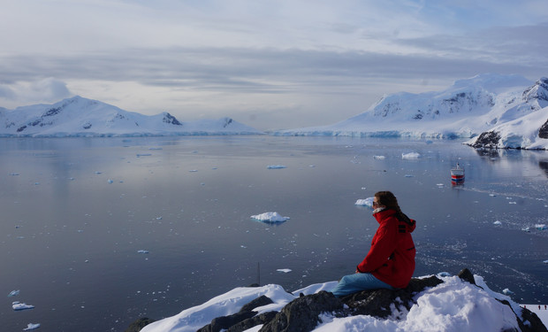 A person in a red jacket sits on a snowy cliff, gazing at an icy landscape with scattered icebergs and a distant ship under a cloudy sky.