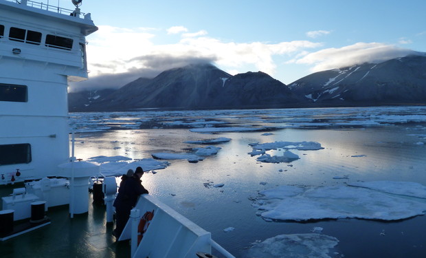A ship moves through icy waters, surrounded by floating icebergs. Two people stand on deck observing the landscape, with distant snow-capped mountains under a clear sky.