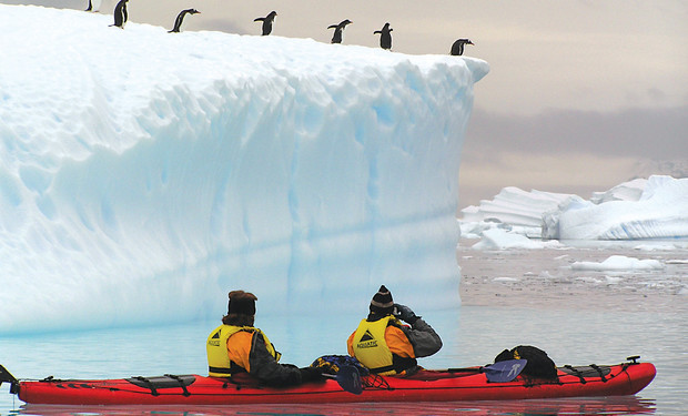 A red kayak with two people in yellow jackets floats on icy water, observing penguins standing atop a large, snow-covered iceberg.