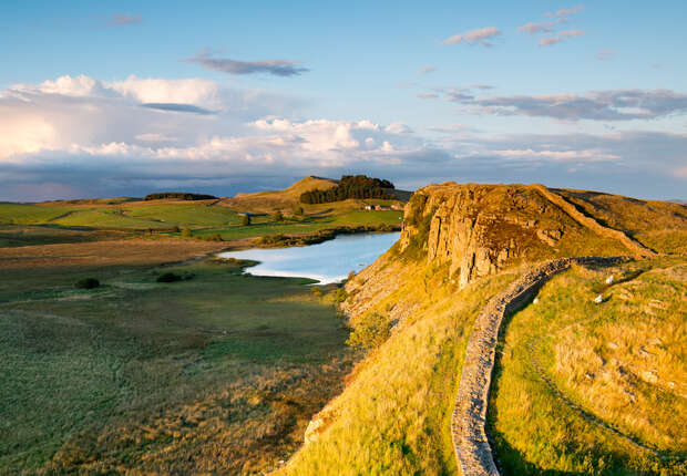 A long, winding stone wall travels along a grassy hill overlooking a serene lake, with rolling green fields extending into the distance under a partly cloudy blue sky.