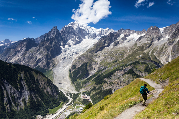 A hiker descends a narrow mountain trail with trekking poles, surrounded by towering peaks and a glacier under a clear blue sky, highlighting an expansive alpine landscape.