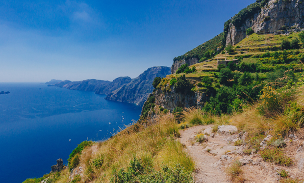 A rugged coastal path winds through grassy terrain, bordered by steep cliffs, with a deep blue sea extending to the horizon under a clear blue sky.