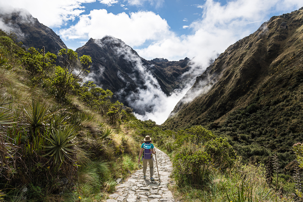 A hiker walks along a stone path, surrounded by lush greenery and steep mountains. Mist drifts through the valley beneath a partly cloudy sky, creating a dramatic, serene scene.