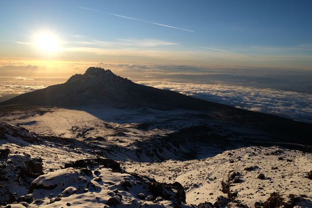 Snow-covered rocky terrain stretches into the distance, leading to a jagged mountain range silhouetted by a setting sun, casting long shadows under a clear blue sky.