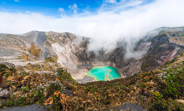 A turquoise crater lake emits steam amidst rocky volcanic terrain, surrounded by lush vegetation and low clouds under a bright blue sky.