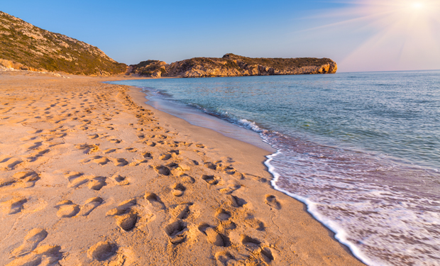 Footprints trail along a sandy beach, leading toward gentle ocean waves under a sunlit sky. Rocky cliffs in the background frame the serene coastal landscape.