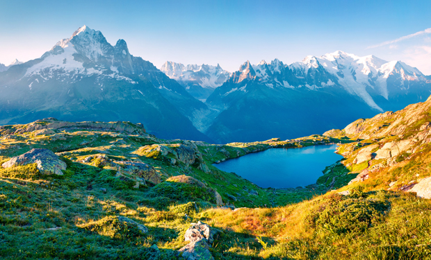 A serene blue lake reflects sunlight, surrounded by rugged green hills. Snow-capped mountains loom majestically in the background under a clear blue sky, creating a tranquil alpine landscape.