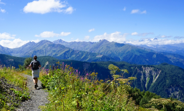 A hiker walks along a narrow, rocky trail, surrounded by green vegetation, with a backdrop of expansive, cloud-tipped mountains under a clear blue sky.
