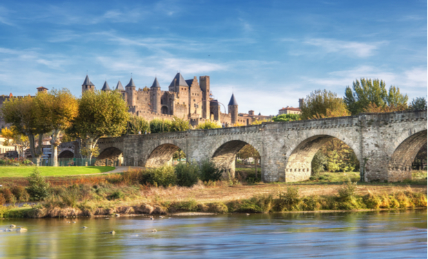 A medieval castle with pointed turrets stands behind an old stone bridge with multiple arches, spanning a tranquil river, surrounded by lush trees under a clear blue sky.