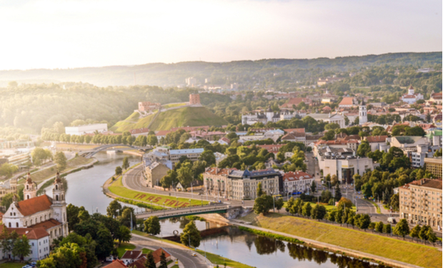 The cityscape features historic buildings under warm sunlight, with a river curving through the landscape, surrounded by greenery. A hillside with ancient structures sits prominently in the distance.