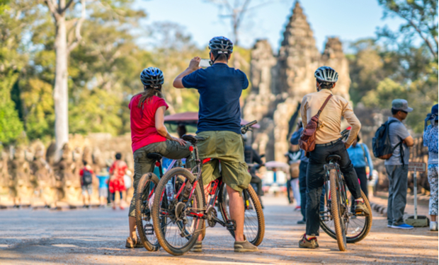 Three cyclists stand with their bikes, taking a photo. They face an ancient stone gate surrounded by tourists in a park-like setting with trees and a clear sky.