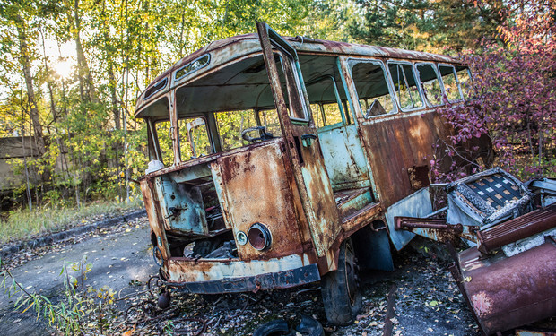 Rusty, abandoned bus leaning to the side, surrounded by dense foliage and overgrown plants, amidst a forest setting. Deterioration suggests long-term neglect, highlighting nature's reclamation of manmade objects.