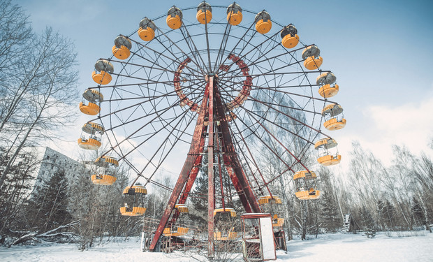 A large Ferris wheel, with yellow gondolas, stands still in a snowy, leafless park. Surrounding bare trees and a distant building enhance the abandoned, wintry atmosphere.