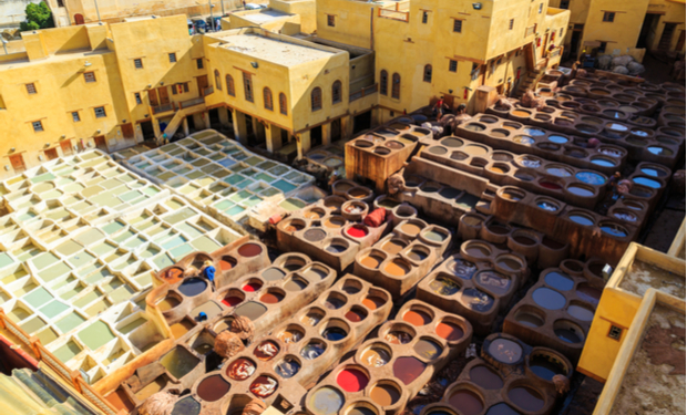 Numerous clay vats filled with colorful dye solutions are arranged in a traditional tannery courtyard, surrounded by yellow buildings, where workers prepare leather in the open air.