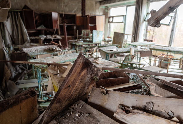 Debris is scattered across a neglected classroom, with overturned desks and chairs amid broken windows, suggesting long-term abandonment and decay. Sunlight filters through the open windows, illuminating the disarray.