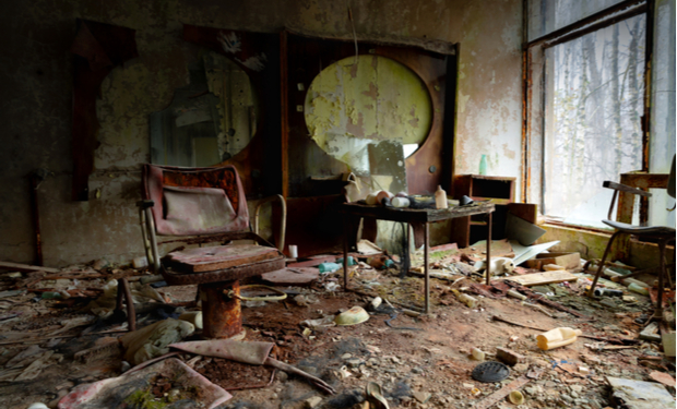 A decayed room with a rusted chair and cluttered table sits amidst debris, peeling paint, and broken mirrors. Sunlight filters through a large, grimy window, highlighting the scene.