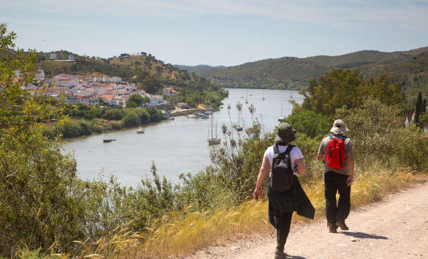 Two people with backpacks walk along a dirt path beside a river, surrounded by lush greenery. In the distance, a small village with white buildings sits on a hillside.
