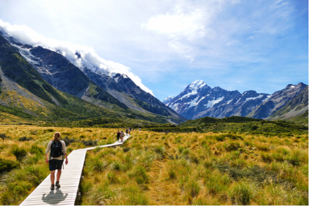A person hikes along a wooden boardwalk surrounded by grassy fields, with snow-capped mountains and a bright blue sky in the background.