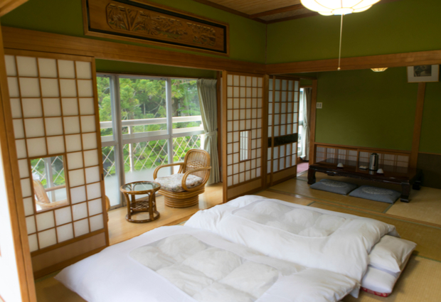 Futons are laid out on tatami mats in a traditional Japanese room with shoji doors. A small sitting area with wicker chairs faces a window overlooking greenery.