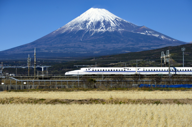 A white bullet train speeds along tracks in the foreground. It passes fields and electrical lines with a snow-capped mountain rising majestically against a clear blue sky in the background.