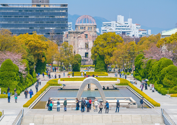 A park features a curved monument over a narrow reflecting pool. People walk nearby, surrounded by manicured trees. In the background, an old structure contrasts with modern buildings.