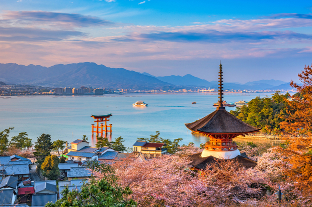 A historic pagoda overlooks cherry blossoms and rooftops near a calm bay, where ferries glide. In the distance, a torii gate stands in the water, framed by mountains.