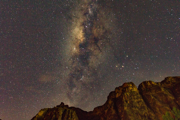 A luminous Milky Way galaxy arcs across a star-filled night sky, illuminating rocky cliffs below in a tranquil desert landscape.