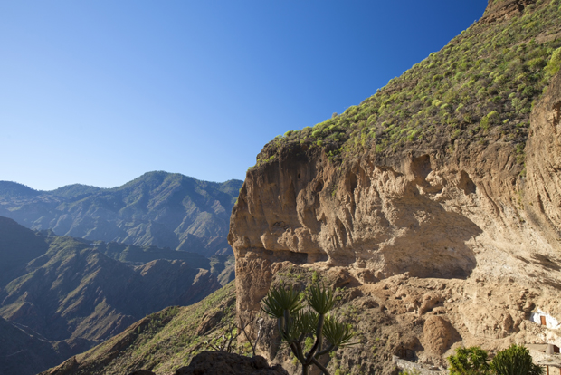 Rugged mountains with steep cliffs extend into the distance under a clear blue sky. Sparse vegetation, including small trees, grows on the arid slopes, creating a stark, natural landscape.
