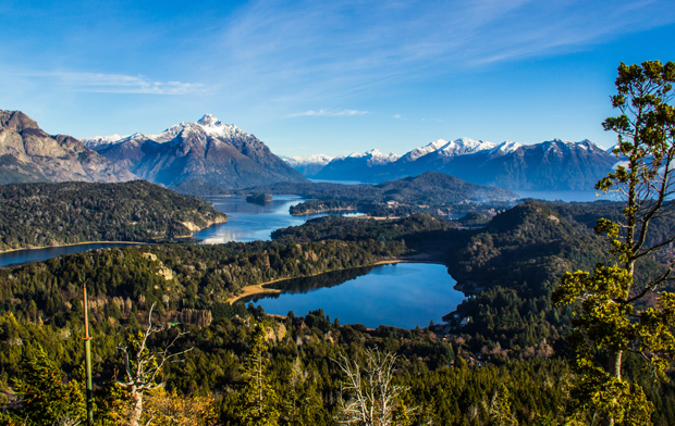 Snow-capped mountains rise in the background, surrounding blue lakes and lush forests. Tall trees frame the foreground, creating a serene, natural landscape under a clear blue sky.