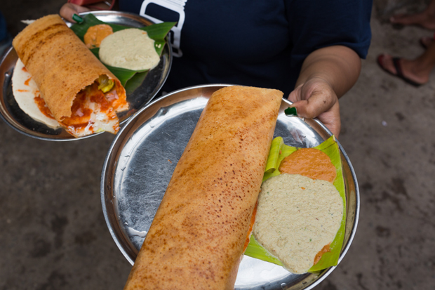 A person holds two metal plates with dosas and chutneys, served on banana leaves. The background shows a rough, outdoor surface and part of another person's foot in sandals.