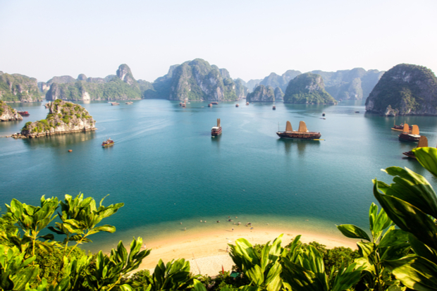 Boats with sails float on a calm turquoise bay, surrounded by towering limestone islands. Lush green foliage frames the foreground, overlooking a sandy beach.