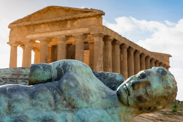 A reclining bronze statue rests in front of an ancient Greek temple with prominent columns, under a blue sky with scattered clouds.
