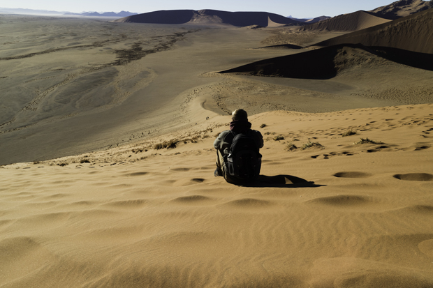 A person with a backpack sits on a sand dune, overlooking an expansive desert landscape with undulating dunes under a clear sky.