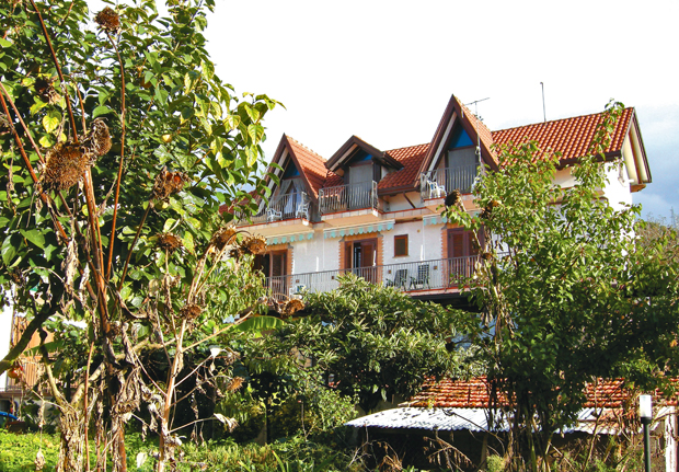 A two-story house with red gabled roofs and balconies is surrounded by green foliage and trees, set against a bright sky.