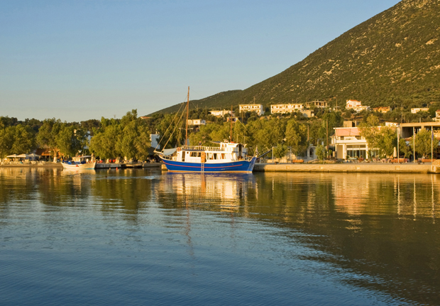 A blue and white boat rests on calm water near a dock. Surrounding it are trees and buildings, with a large hill in the background under a clear blue sky.